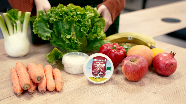 KI generiert: Verschiedene frische Lebensmittel auf einem Tisch: Salat, Joghurt, Karotten, Fenchel, Paprika, Obst. Text: "Naturkost Joghurt".