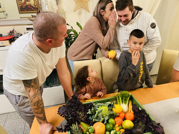 KI generiert: Menschen um einen Tisch mit Gemüse und Obst versammelt, eine Frau flüstert einem Mann etwas zu.