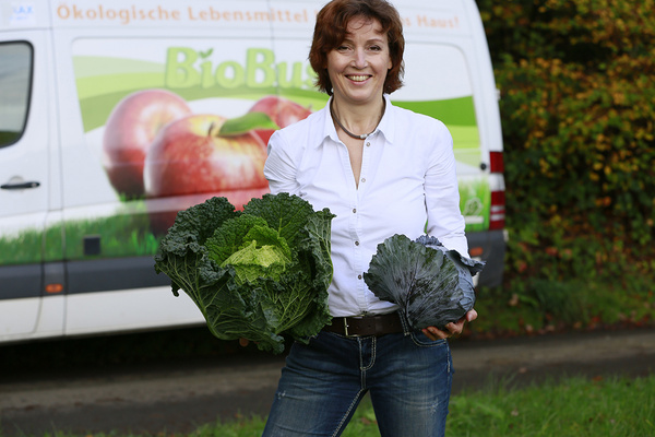 KI generiert: Eine Frau hält einen Korb mit Kürbissen in einem ländlichen Garten mit herbstlicher Kulisse. Neben ihr steht eine Ziege und verschiedene andere Gemüsesorten sind sichtbar.