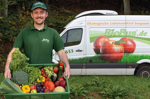 KI generiert: Ein Mann hält eine Kiste mit Gemüse; im Hintergrund ein Lieferwagen mit "BioBus.de"-Aufschrift.