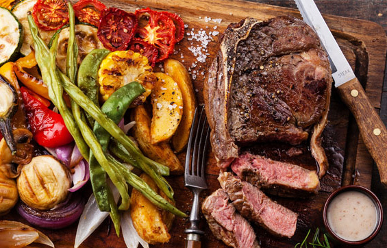 KI generiert: Gegrilltes Steak mit Gemüse und Kartoffeln auf einem Holzbrett, daneben ein Messer und eine Sauce.