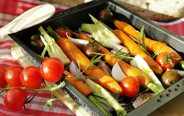 KI generiert: Frisches Gemüse, darunter Karotten und Tomaten, in einer schwarzen Auflaufform auf kariertem Tischtuch.