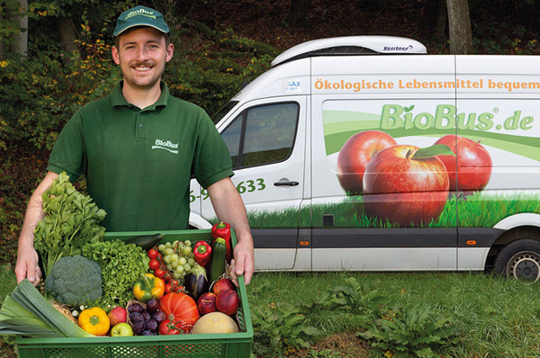 KI generiert: Ein Mann hält eine Gemüsekiste, im Hintergrund ein Lieferwagen mit "BioBus.de". Text: "Ökologische Lebensmittel bequem".